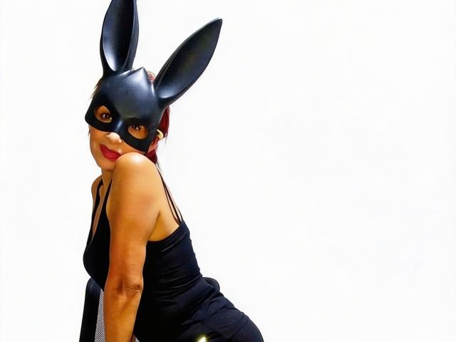 RebecaMonnique wears a black bunny mask with large ears, red lipstick, and a black sleeveless dress. She's looking over her shoulder with a playful expression. Her red hair is visible, and she's positioned slightly to the left. The white background makes her figure stand out. Her dress has a shiny, textured finish, and the mask has a smooth, matte surface. Her right arm is raised, adding to her playful posture. The image has a clear, sharp quality.