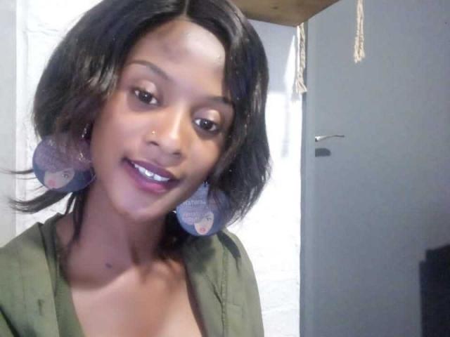 ElsieMeg with short black hair, dark skin, purple lipstick, green shirt, and large purple earrings, smiling at the camera. Boobs partially visible. Sheer nose piercing. Bedroom setting.