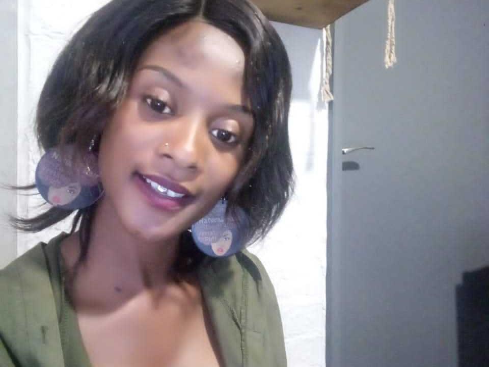 ElsieMeg with short black hair, dark skin, purple lipstick, green shirt, and large purple earrings, smiling at the camera. Boobs partially visible. Sheer nose piercing. Bedroom setting.