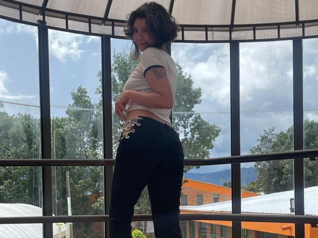 DarckVelvet stands in a glass-enclosed balcony, facing sideways with a slight glance over her shoulder. She has dark, wavy hair and a tattoo on her left arm. She wears a white crop top and black pants with decorative laces on the side. The balcony has a curved, translucent roof. Outside, green trees and a distant mountain are visible. The image has a natural, relaxed composition with a focus on DarckVelvet's upper body and back.