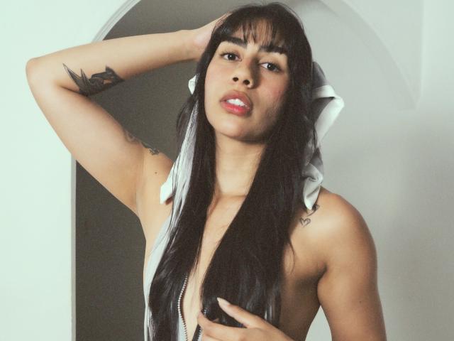 JessicaSoler with long black hair, white bow, and tattoos, holding her tits, hand in hair, looking at camera, pink lips, light skin, and a sexy, confident expression.