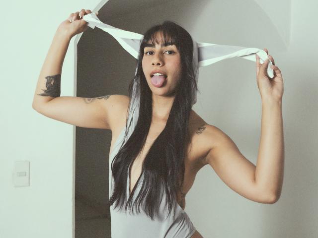 JessicaSoler, a young woman with long black hair and bangs, holds a white piece of fabric stretched above her head with both hands. She has a tattoo on her right upper arm and another on her left shoulder. She sticks out her tongue playfully. She wears a deep V-neck gray dress that exposes her sides and has a muscular build. The image has a soft, slightly grainy texture. Her expression is confident and playful. She is in a white-walled room with a door in the background.