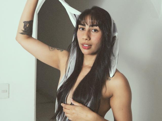 JessicaSoler with black hair, removing a white shirt, exposing her tits, holding the shirt above her head, tattoos on her arm, looking at the camera, soft lighting.