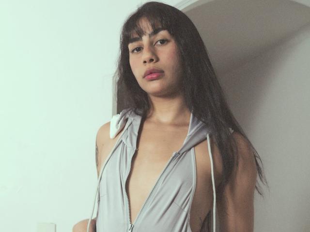 JessicaSoler is in a photograph with a neutral background. She has long, straight black hair with bangs and wears a gray, sleeveless, deep-v zipper dress. Her dress is loose with white drawstrings. She has a light brown skin tone, pink lipstick, and a slightly serious expression. Her small breasts are visible through the zipper opening. The image has a soft, muted color palette. Her tattoo on the right shoulder is partially visible. The overall style is casual and modern.