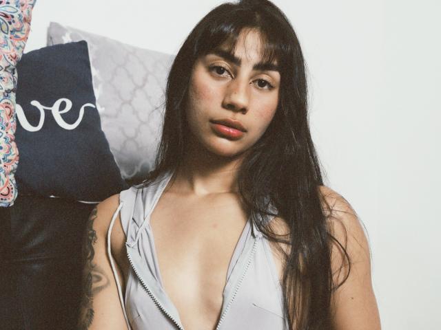 JessicaSoler with long black hair, wearing a low-cut gray top, showing her cleavage, sitting on a black leather couch, looking slightly to the side with a neutral expression.