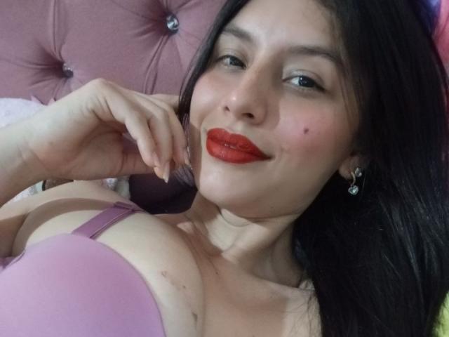 MadissonFontaine with red lipstick, black hair, and purple bra, showing off her tits, lying on pink tufted headboard, smiling, hand near chin, wearing earrings.
