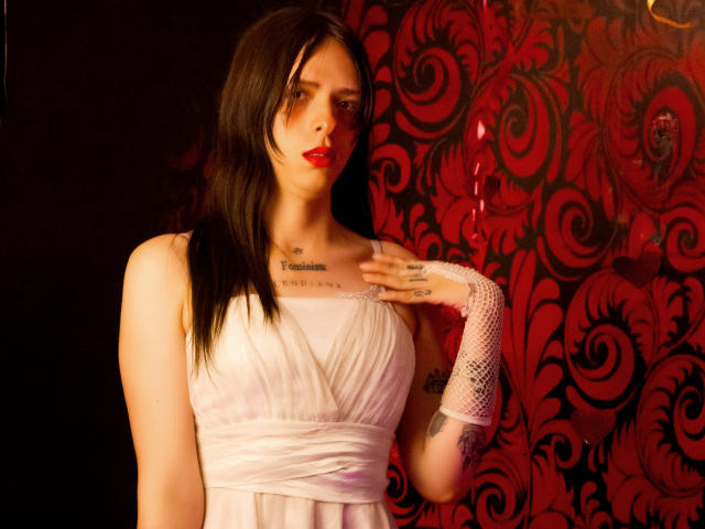 SashaSexiest stands in front of a red and black patterned background. She has long, straight black hair and wears a white, sleeveless, V-neck dress with a gathered waist. Her right hand, adorned with a white fishnet glove, rests on her chest, showcasing tattoos on her hand and chest. She has red lipstick, and her expression is neutral. The lighting is warm, highlighting her pale skin. She has a tattoo below her collarbone that reads "I am not a woman." The image is a photograph with a realistic style.