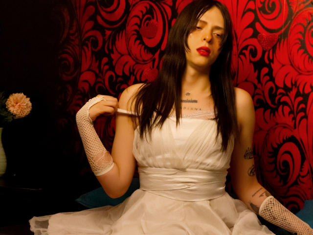 SashaSexiest sits in a white dress with a lace trim, holding a strap. She has long black hair, red lipstick, and tattoos on her chest and arms. She wears white fishnet gloves. The red, black floral patterned background contrasts with her fair skin and blue pillow. A small vase with a pink flower is on the left. The lighting highlights her facial features and the texture of her dress. She has a confident, slightly seductive expression.