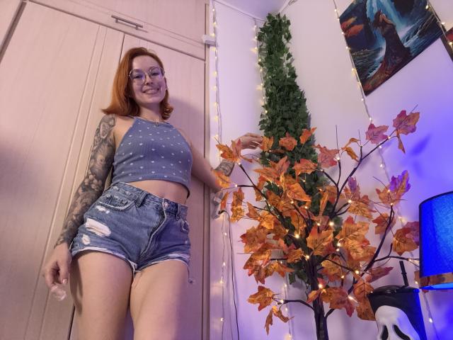 SarahRousen stands in a room, wearing a blue, polka-dotted crop top and distressed denim shorts. She has red hair, glasses, and a tattooed arm. She's smiling, looking down at the camera. A leafy, autumn-themed plant with orange leaves is on the right, next to a blue lamp. A green tree decoration is in the background, and a painting is on the wall. The lighting is purple-blue.