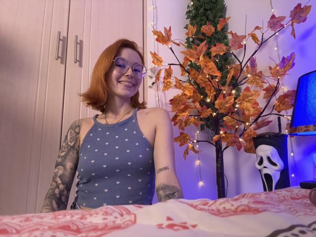 SarahRousen, a red-haired woman with glasses, smiles at the camera. She wears a blue sleeveless top with white star patterns, revealing a tattoo on her right forearm. Her left arm is fully tattooed. She stands in a room with a wooden wardrobe on the left and a decorative autumn tree with orange leaves and string lights on the right. A blue lamp is partially visible behind the tree. The bed in the foreground has a red and white patterned blanket. The image has a casual, homey feel.