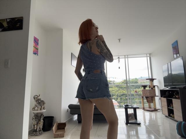 SarahRousen stands in a modern living room with a large window in the background. She has red hair and tattoos on her right arm and upper back. She wears a blue, polka-dotted, sleeveless crop top and short denim shorts. Her right hand is near her chin, and she looks to the left. The room has white walls, a black sofa, a cat tree, and a TV on a stand. A small statue of a bull is on the left. The floor is shiny white tile.