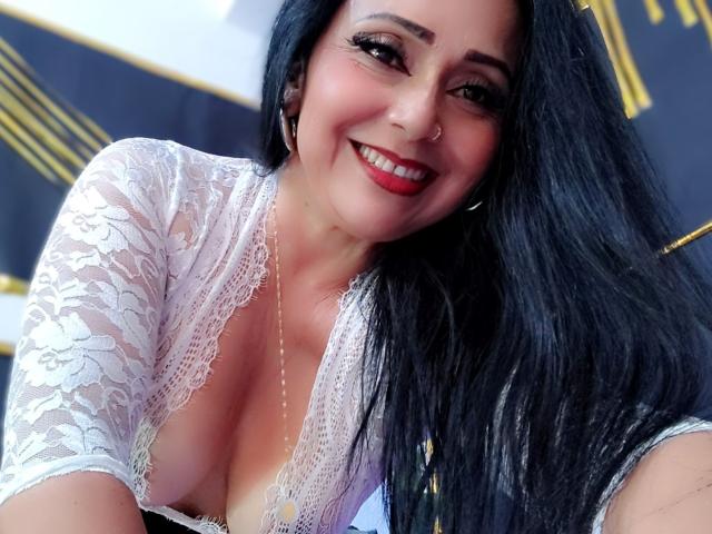 Close-up of Zaria69 with long, straight black hair, smiling with red lipstick. She wears a white lace top with a deep V-neck, revealing cleavage. She has a nose ring in her right nostril and wears small hoop earrings. Her skin is light, and she has dark, well-groomed eyebrows. The lace texture is delicate, contrasting with her smooth skin. The image is brightly lit, highlighting her facial features and hair. The composition focuses on her upper body and face.