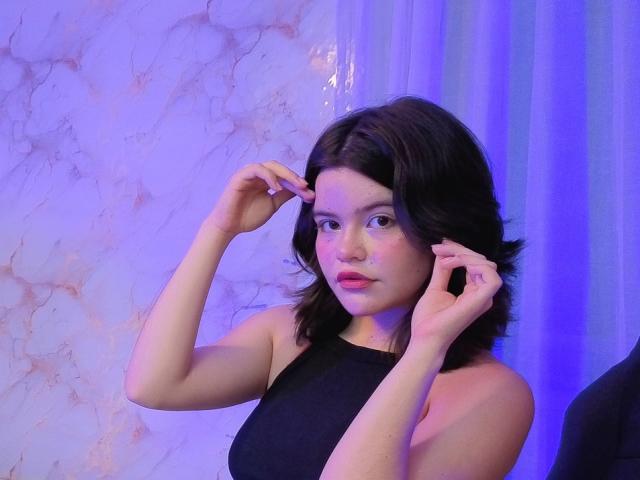 AlexaMissFortune stands in a dimly lit room with a purple-blue light cast on her right. She has medium-length, wavy black hair and is wearing a sleeveless black top. Her hands are near her face, slightly touching her hair. Her expression is neutral, with slightly parted pink lips. The background has a textured, marble-like wall on the left and a semi-transparent curtain on the right. The lighting and her pose create a subtle, introspective mood.
