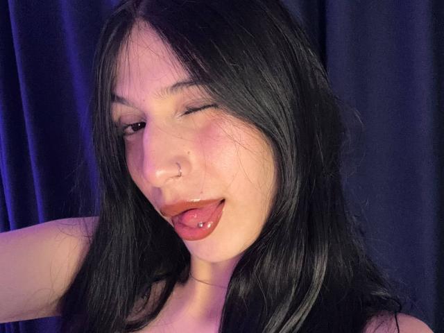 Close-up of SophieBeaumont with dark, straight hair and light skin. She's winking with her right eye, sticking out her red tongue with a silver tongue piercing. She has a small nose ring in her left nostril. Her left arm is raised, partially visible. She wears a black top, and the background is dark blue. The lighting has a purple-blue hue, highlighting her cheek and lip. Her expression is playful and slightly provocative.