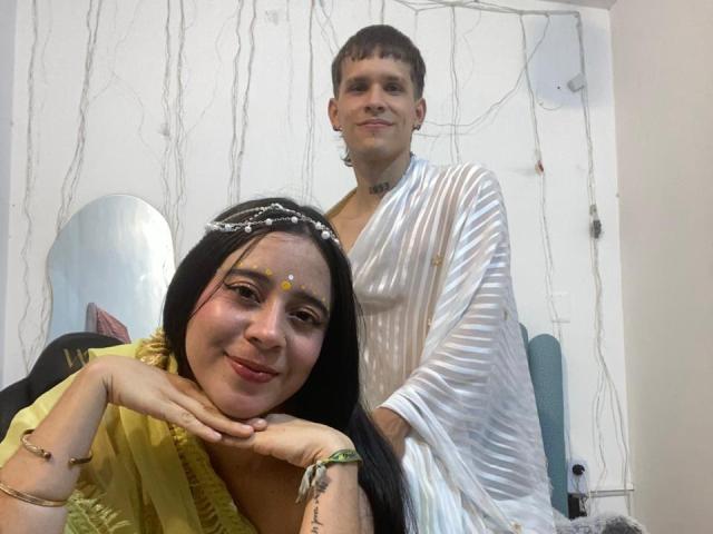 AnjaliRohan with long black hair and a yellow saree rests her head on her hands, smiling. She has yellow dots on her forehead and wears a beaded headband. Behind her, AnjaliRohan, a short-haired person in a translucent white robe, stands with a relaxed posture. The room has white walls with vertical lines and a blue cushion on the right. The image has a casual, informal style with natural lighting.