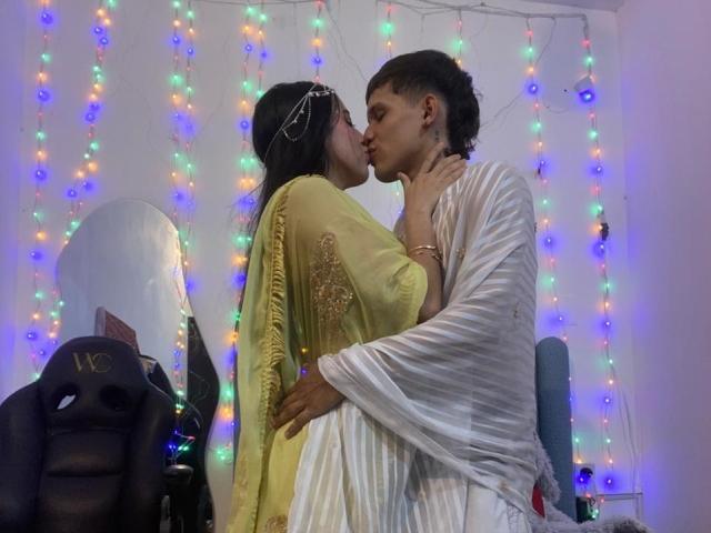 AnjaliRohan and another person kiss in a dimly lit room. AnjaliRohan, wearing a yellow dress with gold embroidery and a silver headpiece, holds the other person's neck. The other person, in a white striped dress, has short, curly hair and a tattoo on their cheek. Multicolored fairy lights hang in the background. An office chair with "MC" on it is on the left. The image is a photograph with a casual, intimate composition.
