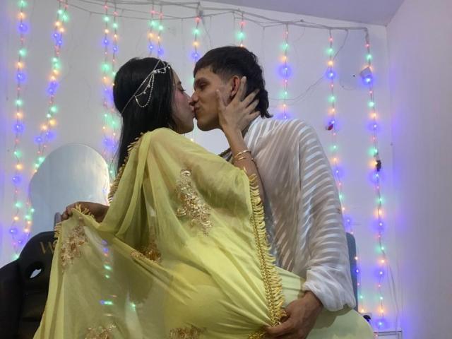 AnjaliRohan, a woman with long black hair, wearing a yellow saree with gold embroidery and a head chain, kisses a man with dark hair, dressed in a white striped shirt. She holds his face with one hand while he holds her with the other. Multicolored string lights hang in the background. Her saree drapes over his lap. The man's right hand is on her waist. The image has a casual, intimate vibe.