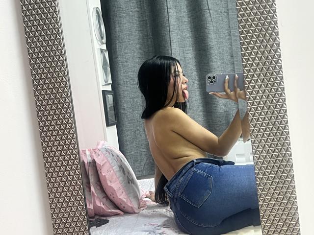 EstrellaXHot, topless, with black hair, sticking out tongue, taking mirror selfie, wearing blue jeans, showing her back and butt, holding phone in patterned mirror.