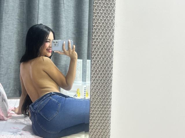EstrellaXHot with long black hair, topless, showing back and ass in blue jeans, taking a mirror selfie with a smiling face, holding a silver phone.
