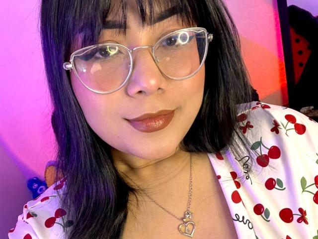 AlexaKiim with black hair, glasses, and red lipstick, wearing a cherry print top, heart necklace, smiling at the camera, pink and purple lighting in the background.