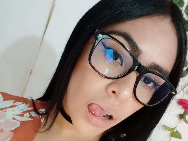 Close-up selfie of StellaBeckker with black straight hair, wearing black-framed glasses reflecting blue light. She has light skin, full pink lips with a small piercing on the lower lip, and is sticking her tongue out slightly. She wears a floral orange top with white patterns and a black choker. The background is blurred with a hint of floral decoration on the right. The image is sharply focused on her face, emphasizing her glasses and expression.