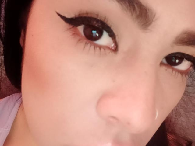 Close-up of StellaBeckker's face. She has medium brown skin, dark eyebrows, and thick black eyeliner wings extending from her upper eyelids. Her brown eyes are slightly open, looking to the right. Her nose is centered in the frame, and her lips are slightly parted. She wears a light purple garment, visible at the bottom edge. The image has a warm, pinkish hue. Her hair is dark and partially visible. The texture of her skin is smooth.