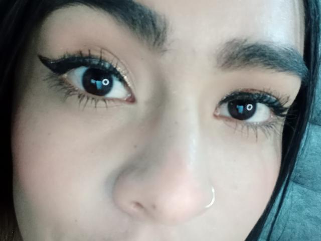 Close-up of StellaBeckker's face, focusing on her eyes. She has dark brown eyes with noticeable reflection in the camera. Her thick, dark eyebrows contrast with her light skin. She wears black eyeliner with a subtle wing, and her long eyelashes are prominent. A small, silver nose ring is on her right nostril. Her straight, dark brown hair is partially visible on the sides. The image has a slightly soft, natural light. The texture of her skin is smooth with a few small freckles.