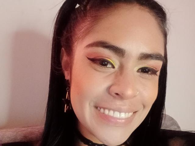 Close-up of StellaBeckker with a light brown complexion and straight black hair in a high ponytail. She has thick, dark eyebrows, yellow eyeshadow with black winged eyeliner, and a small nose ring. She's smiling with white teeth, wearing a black choker and small gold earrings. Soft pinkish-red light is on her right cheek. The image is slightly blurry.