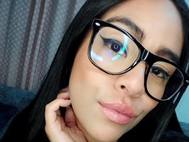 Close-up of StellaBeckker with dark straight hair, black-rimmed glasses reflecting light, and bold black eyeliner. Her skin is smooth and light brown. She has full pink lips and is slightly smiling with a hand near her neck. The image has a clear focus on her face, with the glasses taking up significant space. The light reflection adds a subtle blue glare to her eyes. The texture of her hair is sleek and shiny. The overall composition is centered and tight.