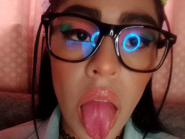 Close-up of StellaBeckker with dark hair, wearing black-framed glasses that have blue and green LED lights reflecting in them. StellaBeckker has dark eyebrows, glossy pink lips, and her tongue is fully extended and slightly shiny. She's wearing a light blue garment and a black choker necklace. The background is pink with a subtle pattern. The image is focused on StellaBeckker's face, highlighting the LED lights and her tongue.