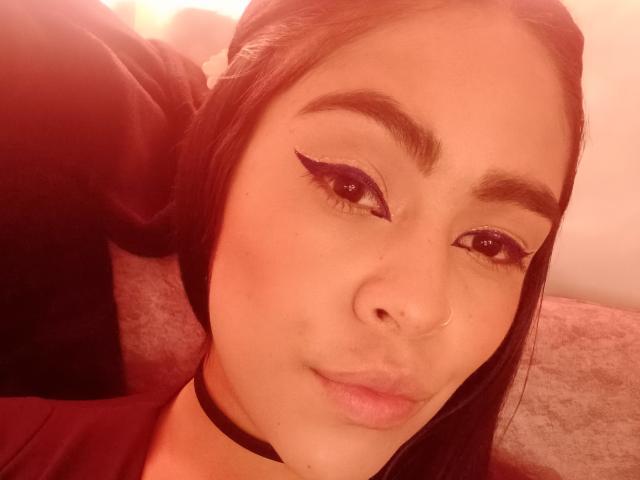 Close-up of StellaBeckker's face with warm, reddish lighting. She has long black hair, dark eyebrows, and wears purple eyeliner with a winged shape. She has a small nose ring in her right nostril and a black choker around her neck. Her lips are slightly parted, and she has a neutral expression. The image has a soft, slightly blurred texture, emphasizing her facial features. The background is indistinct, with a light-colored, textured surface behind her. The overall composition is centered on her face.