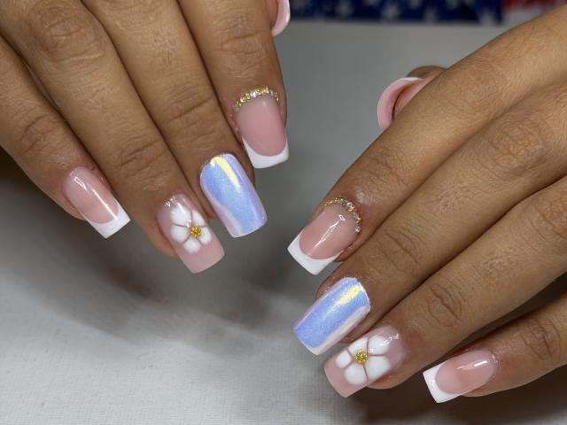 EstrellaXHot's nails are pink with white tips, featuring blue and silver glitter, and small pink flowers with gold centers. Her hands are close together, showing off the manicure.