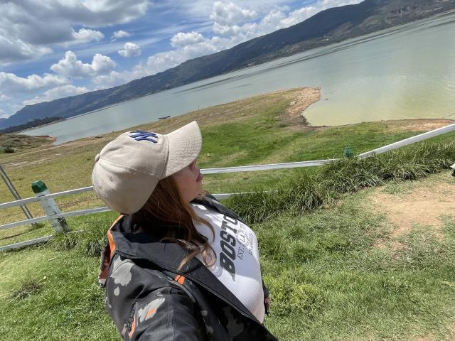 A photograph of JennyLawry stands in a grassy area, facing a lake. She wears a beige cap with a blue logo, a black jacket with orange accents, and a white shirt with black text. Her long brown hair is visible. The lake is greenish, bordered by grass and a wooden fence. The background includes mountains and a cloudy sky. The composition uses a natural, slightly elevated viewpoint, emphasizing the landscape. JennyLawry is positioned in the lower left, looking towards the right.