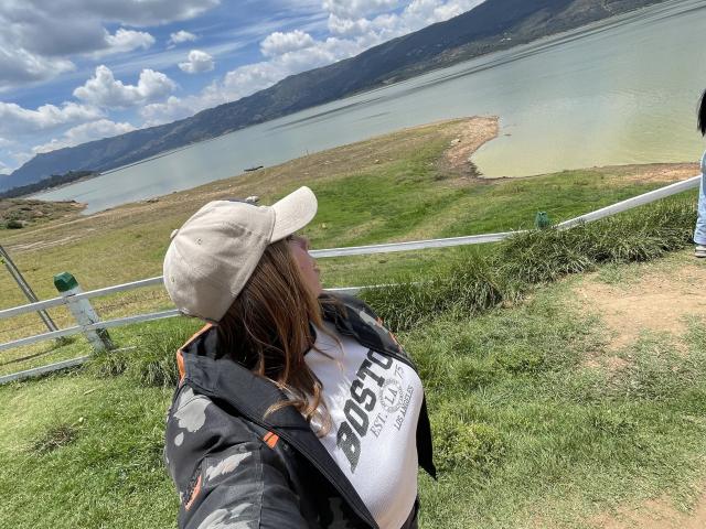 JennyLawry stands on a grassy hill, facing a large, calm lake with a mountain in the background. She wears a beige cap, white "BOSATI" shirt, and black camo jacket. Her brown hair is partially visible. The sky is blue with fluffy clouds. A metal fence runs horizontally behind her. The grass is green and the lake is turquoise. The hill slopes downward to the lake. The composition emphasizes JennyLawry's relaxed posture and the serene landscape. Natural light highlights the scene.