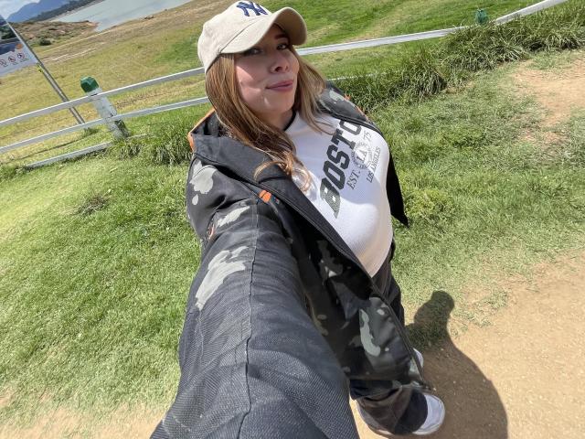 JennyLawry takes a selfie outdoors. She's wearing a beige NY cap, a white "DOSE OF TALENT" shirt, and a black camo jacket. Her brown hair is loose, and she has a slight smile. She extends her left arm towards the camera, creating a small shadow on the grass. The jacket has visible wear, and the grass is green with a dirt path. Her white sneakers are partially visible. The image has natural sunlight.