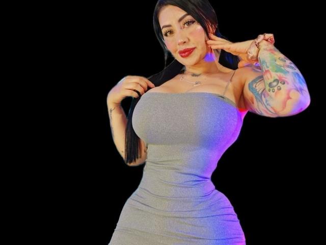 Kkole with large tits, wearing a tight gray strapless dress, has a tattooed arm, black hair, and red lipstick, posing with a hand near her face.