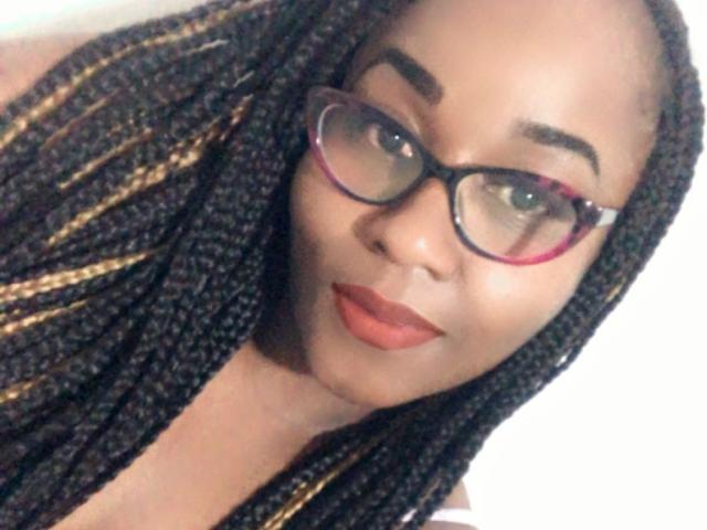 Close-up photo of Yirily with dark brown skin and long, thick, black braids with yellow highlights. Yirily has full, dark eyebrows, green eyes behind black-framed glasses with a red accent, and red lipstick. Yirily's braids hang over the right side of the face. The image is slightly blurred, with a soft, warm light highlighting Yirily's face. Yirily's expression is neutral, with a slight, confident smile. The background is plain and out of focus.