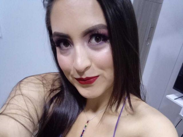 VenusSweetPalmer is a young woman with long, straight brown hair and fair skin. She has dark, heavily made-up eyes with purple eyeshadow, thick eyelashes, and red lipstick. She's wearing a purple halter top and a necklace with small, colorful beads. Her expression is slightly smiling. The photo is a close-up, focusing on her face and upper shoulders. The lighting is even, highlighting her makeup and skin. Her hair is parted in the middle.