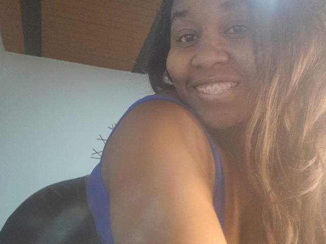 Yirily, a dark-skinned woman with long, wavy brown hair, smiles at the camera. She wears a blue sleeveless top, showing her right shoulder and part of her upper chest. Her teeth are slightly visible, and her right arm is raised. The light source is from the top right, creating a bright spot on her face. She has a relaxed, friendly expression. The background is blurred, with a white wall and wooden ceiling.
