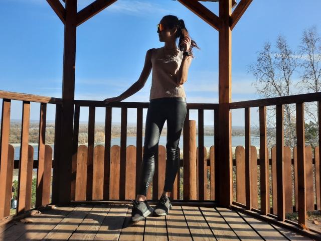 A photograph of MissKinney standing on a wooden balcony. She's wearing a tan sleeveless top, black leggings, and black ankle boots. Her right hand is in her hair, and she's looking to the left. The balcony has vertical wooden slats, and the wooden floor has a grid pattern. Clear blue sky and distant trees are visible. Sunlight casts shadows on the balcony floor. She's centered, with a relaxed, casual posture.