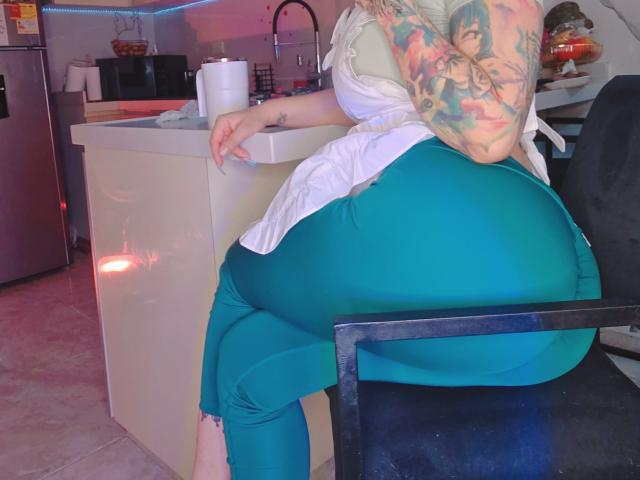 A photo of Kkole, a heavily tattooed woman with teal-green pants and a white sleeveless top. She's sitting on a black chair with her back to the camera, her right arm resting on a kitchen counter. Her large, round butt is prominently displayed. Her tattoos are colorful and detailed, covering her upper arm. The kitchen has a white counter, a stainless steel sink, and a microwave. The floor is tiled in a pinkish hue. The lighting is soft, with a slight pinkish-purple glow.