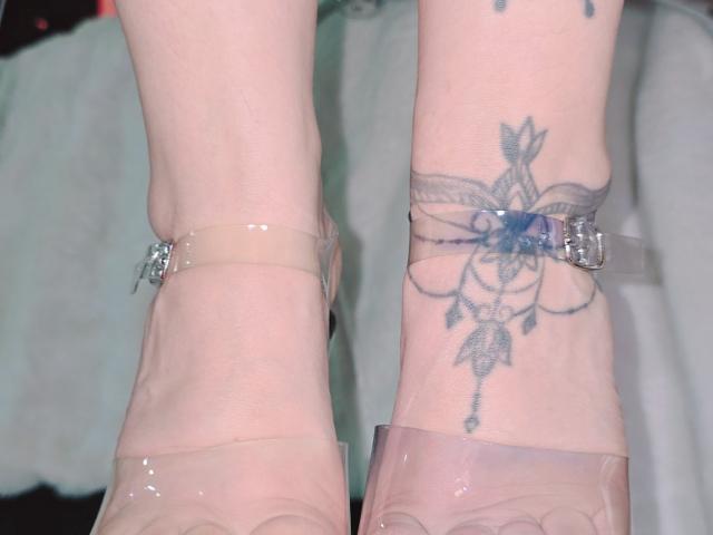 Close-up of Kkole's ankles. Kkole has a tattoo on the right ankle featuring a detailed blue and gray floral design with a central flower and delicate lines extending downward. Kkole's ankles are adorned with clear plastic straps, each with a small silver buckle. The skin is light with visible veins. The tattoo is centered on the ankle, and the straps are slightly glossy, reflecting light. The image focuses on the tattoo and straps, with minimal background detail.