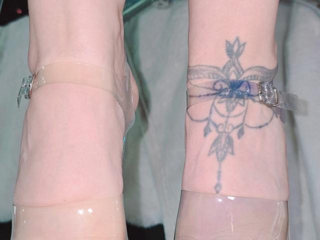 Kkole's pale, tattooed arm with a blue, intricate flower tattoo, wearing clear plastic cuffs on her wrist and a transparent plastic strap around her ankle.
