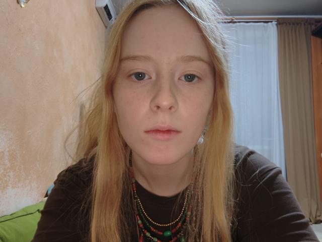 MichaelEuber has pale skin, blue eyes, and blonde hair, wearing a brown shirt with a beaded necklace, looking straight into the camera with a neutral expression.