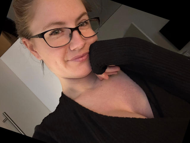 Chantals-passion-hot with glasses, smiling, black ribbed shirt, exposing cleavage, right hand on neck, light skin, blonde hair in a bun, slight pimple on cheek, indoor setting.