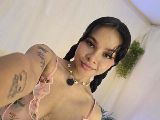 Yurimy with dark hair in braids, pink lace top, tattoos on arm, smiling, necklace with black and white flowers, holding out phone for selfie, light skin, small tits.