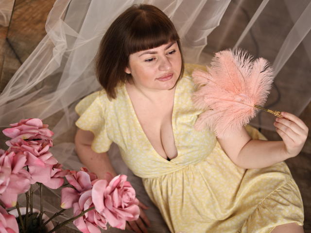 ZannaDreil with short brown hair, wearing a yellow polka-dot dress, holds a pink feather duster, sitting near pink roses, showing her tits, with a gentle smile.
