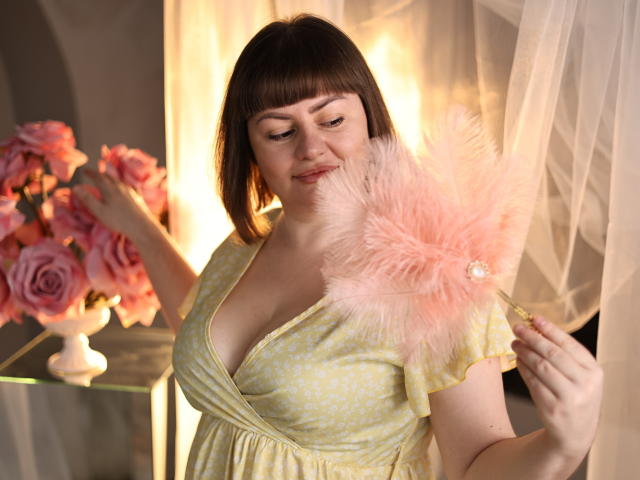 ZannaDreil with short brown hair, pale skin, and a curvy body, holding a pink feather duster, wearing a yellow polka dot dress, smiling, next to a pink rose vase.