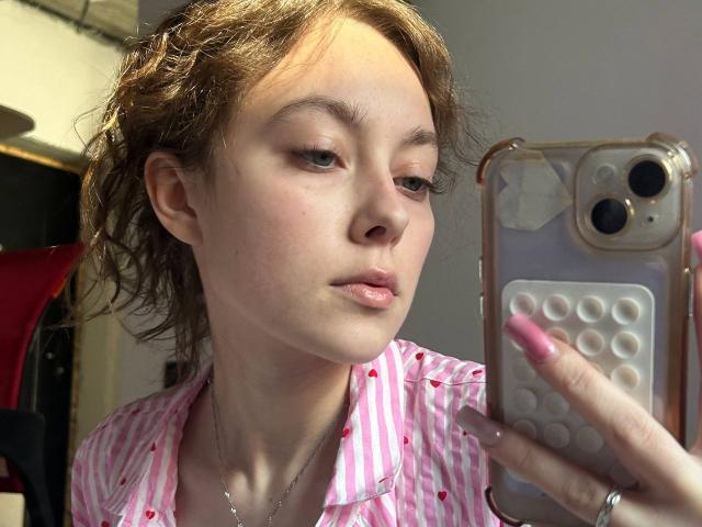 KristalRozon with curly brown hair, pink striped shirt, and pink nails, taking a selfie with a phone, slightly pouty lips, light skin, and subtle makeup.