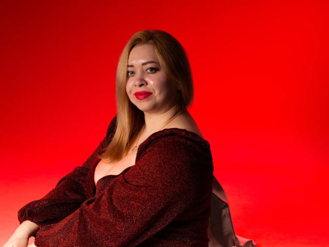 OliviaVerr with red lipstick, smiling, wearing a dark red textured off-shoulder top, light brown hair, red background, slightly turned, showing her upper body and subtle ass.